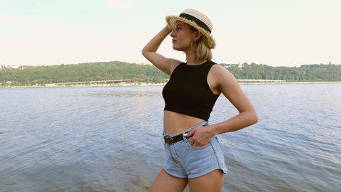 A young woman in short shorts and a black t-shirt in a hat stands on the beach near the river in the city. Slow motion. Relax in nature