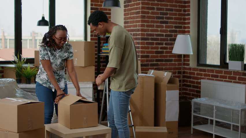 African american couple wrapping cardboard boxes with sticky tape to move in new apartment flat. Packing up furniture and home belongings in packages with adhesive roller for relocation. Royalty-Free Stock Footage #1090301463