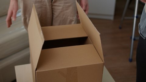 Couple packing up storage things in cardboard boxes, using adhesive sticky tape to wrap package for transportation. Preparing to move in household apartment bought on loan. Close up.