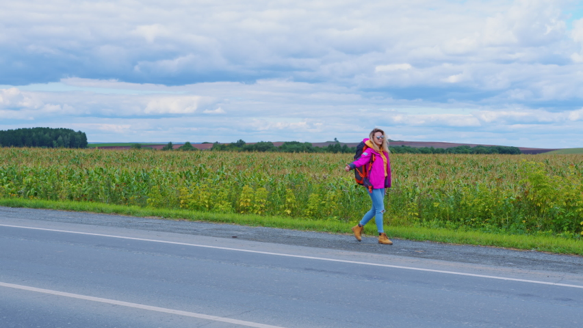 Pretty blonde is slowly walking back along roadside near field trying to stop passing car. Hitchhiker girl with backpack holding raised hand and thumb. Young woman hitches ride near cornfield