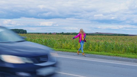 Pretty blonde is slowly walking back along roadside near field trying to stop passing car. Hitchhiker girl with backpack holding raised hand and thumb. Young woman hitches ride near cornfield