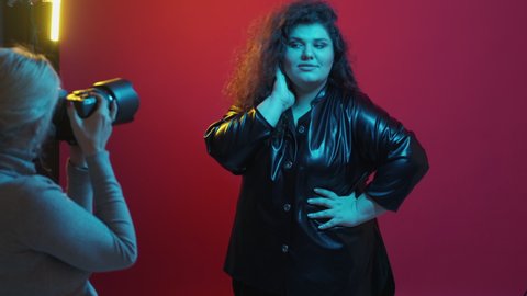Plus size fashion. Professional photo shoot. Body positive. Confident chubby model woman posing for photographer with camera on magenta pink neon light studio background.