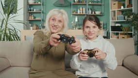 Grandmother and Young Granddaughter Having Fun Together at Home, Sitting on Sofa Holding Gamepads, Playing Racing Video Games. Multigenerational Family Younger Older Use Modern Wireless Tech Concept