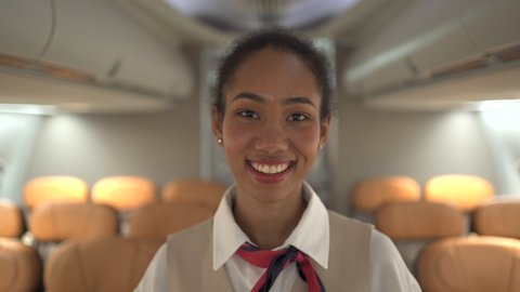Beautiful female airline cabin crew looking and smiling to camera in the cabin. Confident and happy female flight attendee welcoming the passenger onboard.