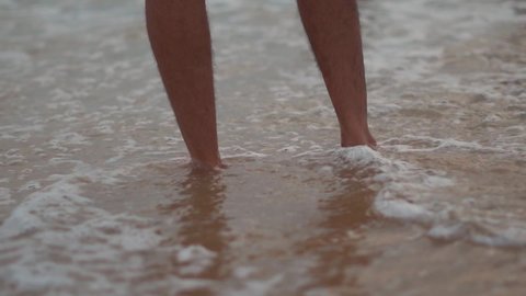 Low angle shot of legs of an Indian man standing in the water of the sea waves of the beach at Baga beach in Goa, India. Man soaks his legs on the beach water. Man enjoying his summer holidays. 