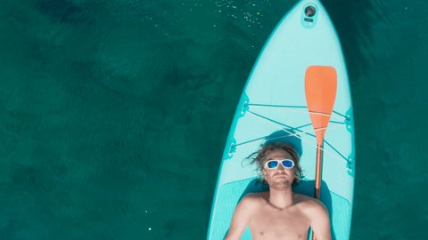 Directly above aerial shot: Man on vacation relaxes lying down on stand up paddle board in turquoise coloured tropical water. Man enjoying summer vacation 