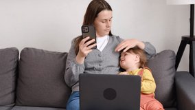 Attractive young mother remote working online sitting on cozy couch near cute little daughter, mom using laptop and smartphone. Modern family use gadgets concept. Virtual home office, distance job