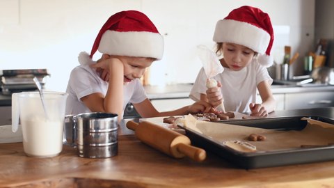In Santa hats. Putting cream. Two children is cooking sweets on the kitchen together.