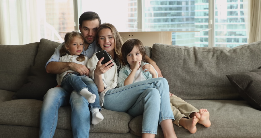 Happy couple with cute little 6s kids spend weekend use smartphone, make video call, watching vlog, enjoy free time on internet. Young generation using modern wireless tech for fun and leisure concept Royalty-Free Stock Footage #1090320149