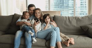 Happy couple with cute little 6s kids spend weekend use smartphone, make video call, watching vlog, enjoy free time on internet. Young generation using modern wireless tech for fun and leisure concept