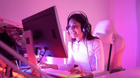 Selective focus of a young pretty Asian transgender gamer wearing a gaming headset sitting on a chair, clicking the keyboard and mouse, enjoyed playing online video games on a computer in a bedroom.