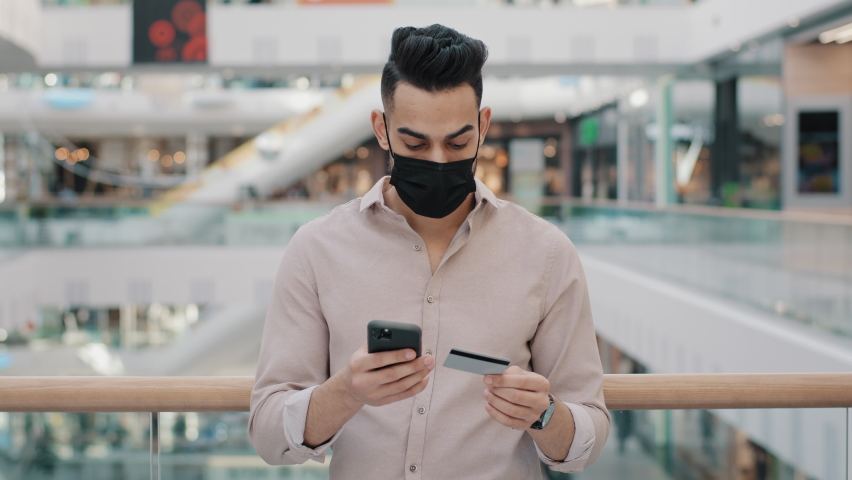 Indian arabian man in medical mask with phone pays online booking mobile service food delivery enters bank card number payment with virtual money shopping with smartphone receives paper bag with order Royalty-Free Stock Footage #1090324825