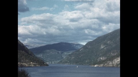 1950s: man holding clapperboard. Waterfall in mountains. Fjord and mountain view. Clouds and blue sky above fjord. Boat enters harbour.