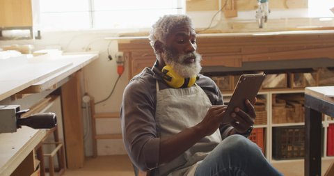 African american male carpenter using digital tablet sitting in a carpentry shop. carpentry, craftsmanship and handwork concept