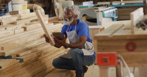 African american male carpenter with digital tablet choosing wooden plank in a carpentry shop. carpentry, craftsmanship and handwork concept
