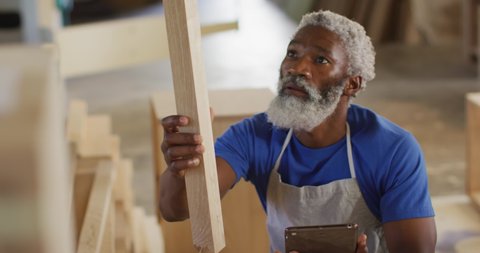 African american male carpenter with digital tablet choosing wooden plank in a carpentry shop. carpentry, craftsmanship and handwork concept