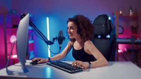 Young woman wearing headphones playing computer game neon fashion room, winner. Female gamer girl blogging, commenting hard match, looking at computer monitor, using computer mouse and keyboard