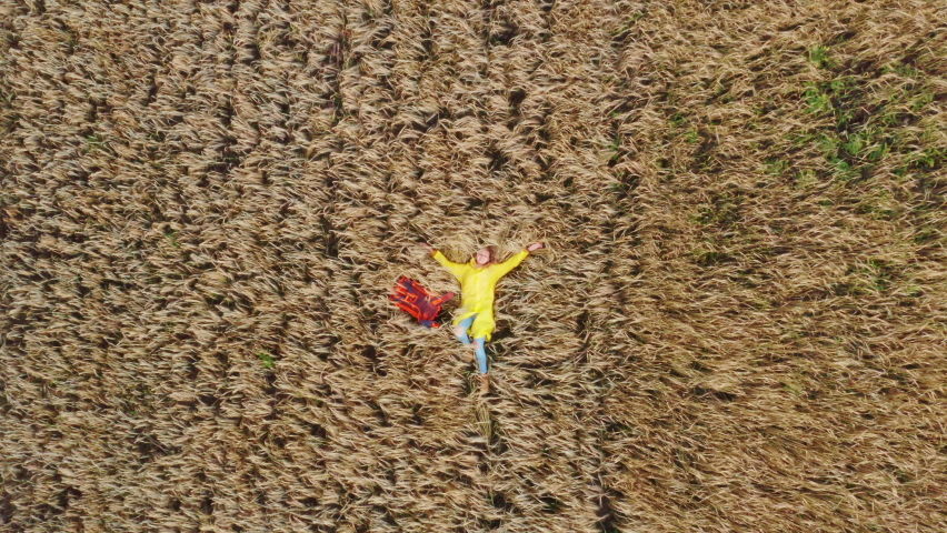 A young girl, a traveler and a hitchhiker, lies next to a backpack on a cereal field sown with wheat or rye. Summer cloudy day. Aerial top view
