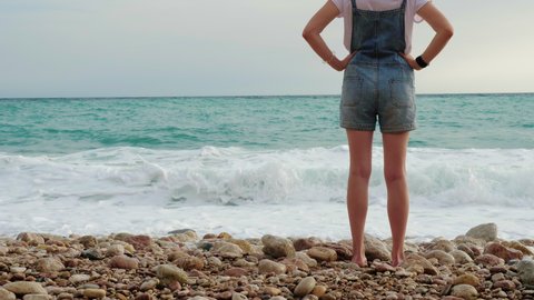 Slender girl in short denim overalls stands on pebbly beach of seashore. Back view. Faceless slender young woman admires marine landscape. Lady spends vacation or holidays on ocean coast