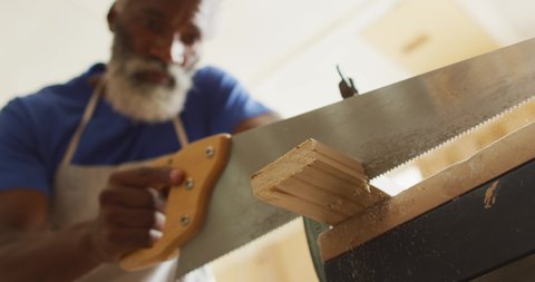 African american male carpenter cutting a wooden plank using a hand saw in a carpentry shop. carpentry, craftsmanship and handwork concept