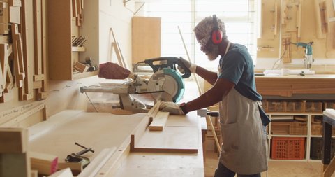 African american male carpenter cutting wooden plank with electric chop saw in a carpentry shop. carpentry, craftsmanship and handwork concept