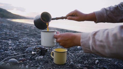 Faceless woman pours coffee from cezve into white cup sitting on pebble beach against endless sea with morning sunlight reflections, detail view hands holds mug, hiker have breakfast on seaside