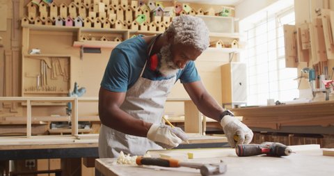 African american male carpenter making pencil markings on wooden plank at a carpentry shop. carpentry, craftsmanship and handwork concept