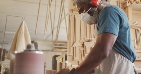 African american male carpenter wearing face mask using belt sander machine to polish wood. carpentry, craftsmanship and handwork concept