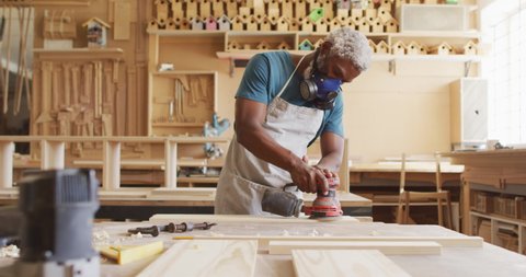 African american male carpenter wearing protective mask sanding a wooden plank with electric sander. carpentry, craftsmanship and handwork concept