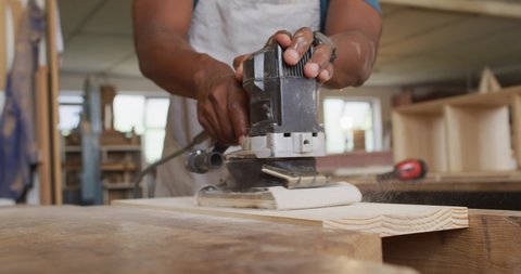 Mid section of african american male carpenter using an electric grinder to grind wooden plank. carpentry, craftsmanship and handwork concept