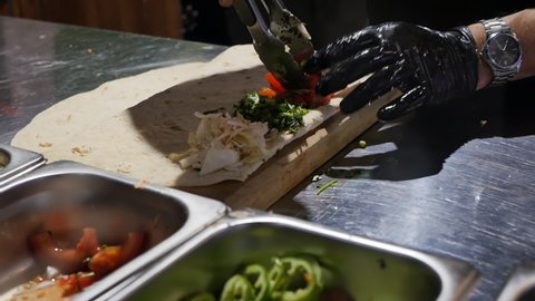 the hand of the cook in a black glove puts fresh pieces of tomato with culinary tongs into a thin pita bread with other vegetables. The process of making lula kebab and shish kebab.