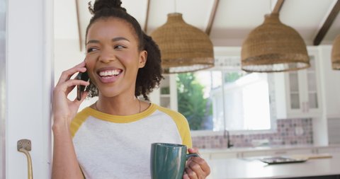 African american woman holding coffee cup talking on smartphone at home. lifestyle and technology concept