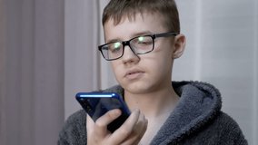 Sad Child in Glasses Holds a Smartphone in Hands, Talks via Video Link in Room. Tired smart boy communicates online through app on a mobile phone. Reflection of glare in glasses. Internet, education.