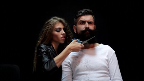 Cutting long beard. Woman hairdresser does the beard to a brutal man in the salon. Barber scissors and straight razor, shaving. Creative advertising, woman shaving.