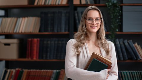 Portrait smiling librarian teacher science professor woman in eyeglasses hold books stack posing at library. Happy clever beauty female literature lover enjoy literary hobby textbooks at bookshelves 
