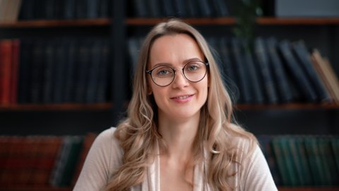Closeup portrait smiling curly blonde woman in vintage eyeglasses posing at library bookshelves with positive emotion. Happy beautiful female face science professor librarian literature lover teacher 