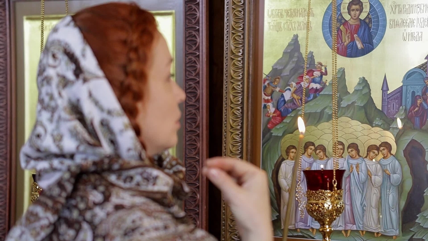 a young woman in a headscarf prays in an Orthodox church and puts candles in front of icons Royalty-Free Stock Footage #1090340963