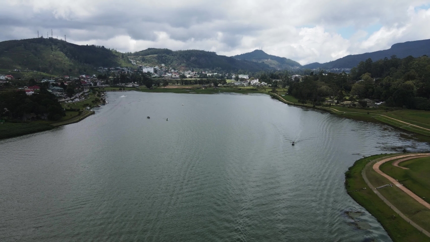 Water sports enthusiasts sail across the large reservoir Gregory Lake in the heart of the Hill City tea region in Sri Lanka between the mountains on a cloudy day. Backwards drone lifting dolley shot