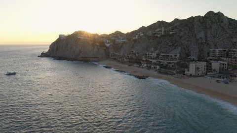 Drone shot of resorts on Playa El Médano with mountains in the distance in Cabo San Lucas Mexico, starting on the ocean then revealing the buildings