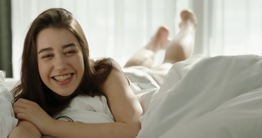 Pretty asian gen z girl looking at camera and smiling while kidding around in cozy bed early in morning. Cute girl relaxing in sunshine 4k portrait footage