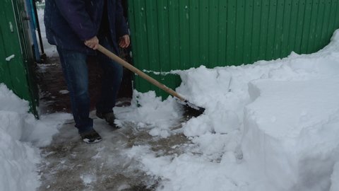 A man in a jacket and jeans is shoveling snow in front of the yard. Clearing the area in winter after a snow storm. Green fence in the background. Snow falls from the sky