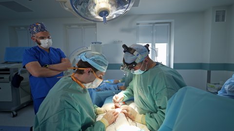 KYIV, UKRAINE - August 2021: Male surgeons sit at both sides from a patient operating his arm. Anesthesiologist standing beside controlling the patient's condition.