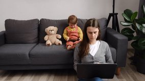 Virtual home office, distance job. Young mother remote working online sitting on warm floor at home using laptop, pretty little daughter child holding smartphone. Modern family use gadgets concept