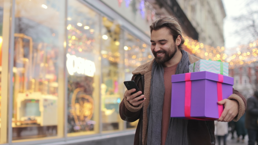 Joyful young man in warm jacket walking on city street with shining garlands around, carrying gift boxes and using modern smartphone. Happy bearded man enjoying shopping for winter holidays. Royalty-Free Stock Footage #1090354113