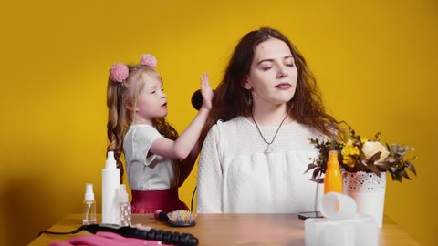 A little girl hairdresser dries her client's hair on a yellow background. Children as adults