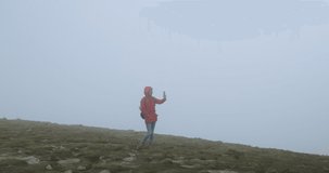 girl tourist with a backpack on the edge of a mountain among fog and clouds, holds a smartphone in her hands, making a selfie or video call. Zero visibility, wide shot, Carpathians, Ukraine, Europe.