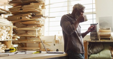 African american male carpenter using smartphone while drinking coffee in a carpentry shop. carpentry, craftsmanship and handwork concept