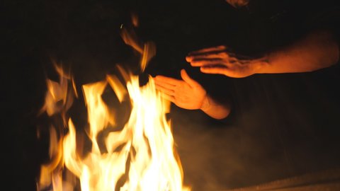 Low view of young tourist rubs his hands in front of an open bonfire from mangal at night. Man warms his arms by a fire from metal brazier against the black background. Concept of warmth and tranquil