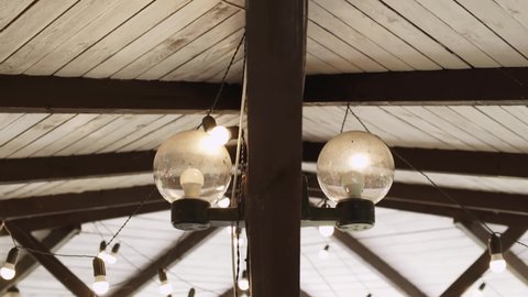 Round glass old lamps with energy saving LED bulbs on wooden pole under roof of the building. The ceiling is decorated with garlands of light bulbs hanging from wires. Countryside lighting.