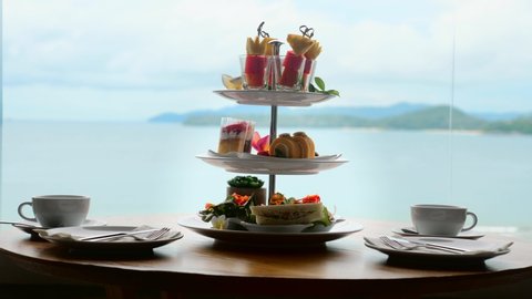 High tea set with assortment of sweets, cakes, sandwiches, fruit canapes on table. Afternoon tea stand with sweet treats, cups for two persons. Tropical sea and mountains landscape background.